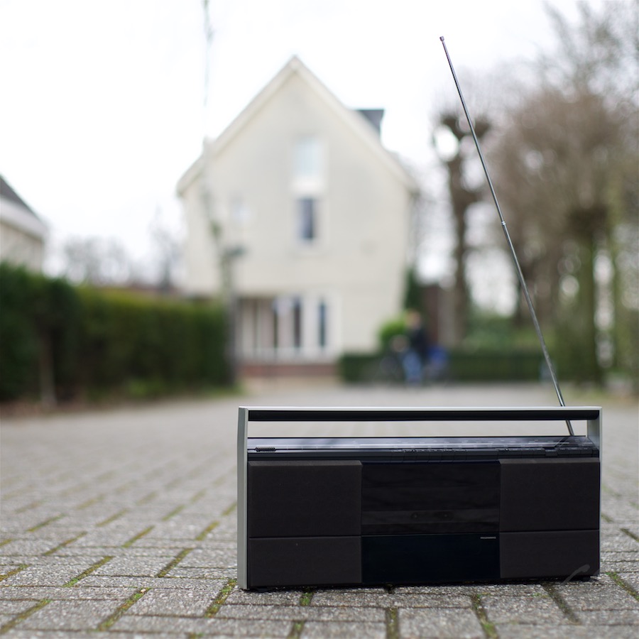 Radio is een vriendje. Foto van een transistorradio op een klinkerweg. Op de achtergrond een onscherp huis.