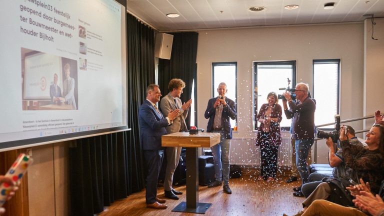 Feestelijke opening van mediaplatform Nieuwsplein33 in Amersfoort. Met confetti, een beamerscherm en mensen achter een statafel.